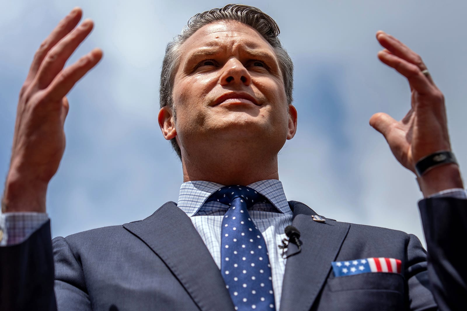 A close up shot of Pet Hegseth in a navy blue suit. His hands are raised up as if in worship.